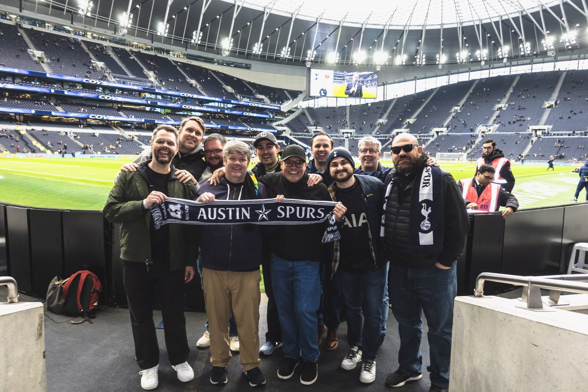 Austin Spurs members at the front of the stand inside Tottenham Hotspur Stadium, holding an Austin Spurs scarf along the front row before kickoff.