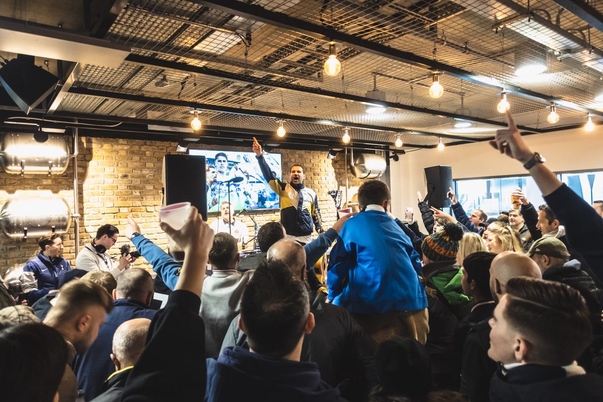 A packed bar before a match, drinks and arms in the air while someone leads a song from the front.