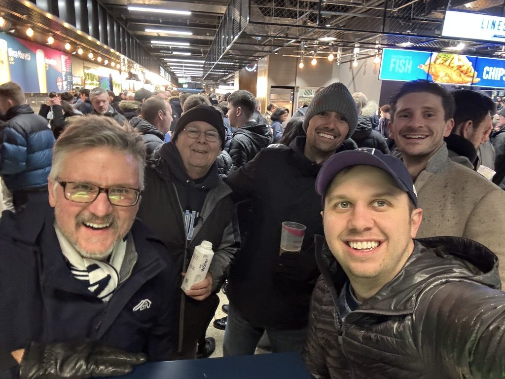 Five supporters leaning in for a selfie on the stadium concourse, food stalls and a queue behind them.