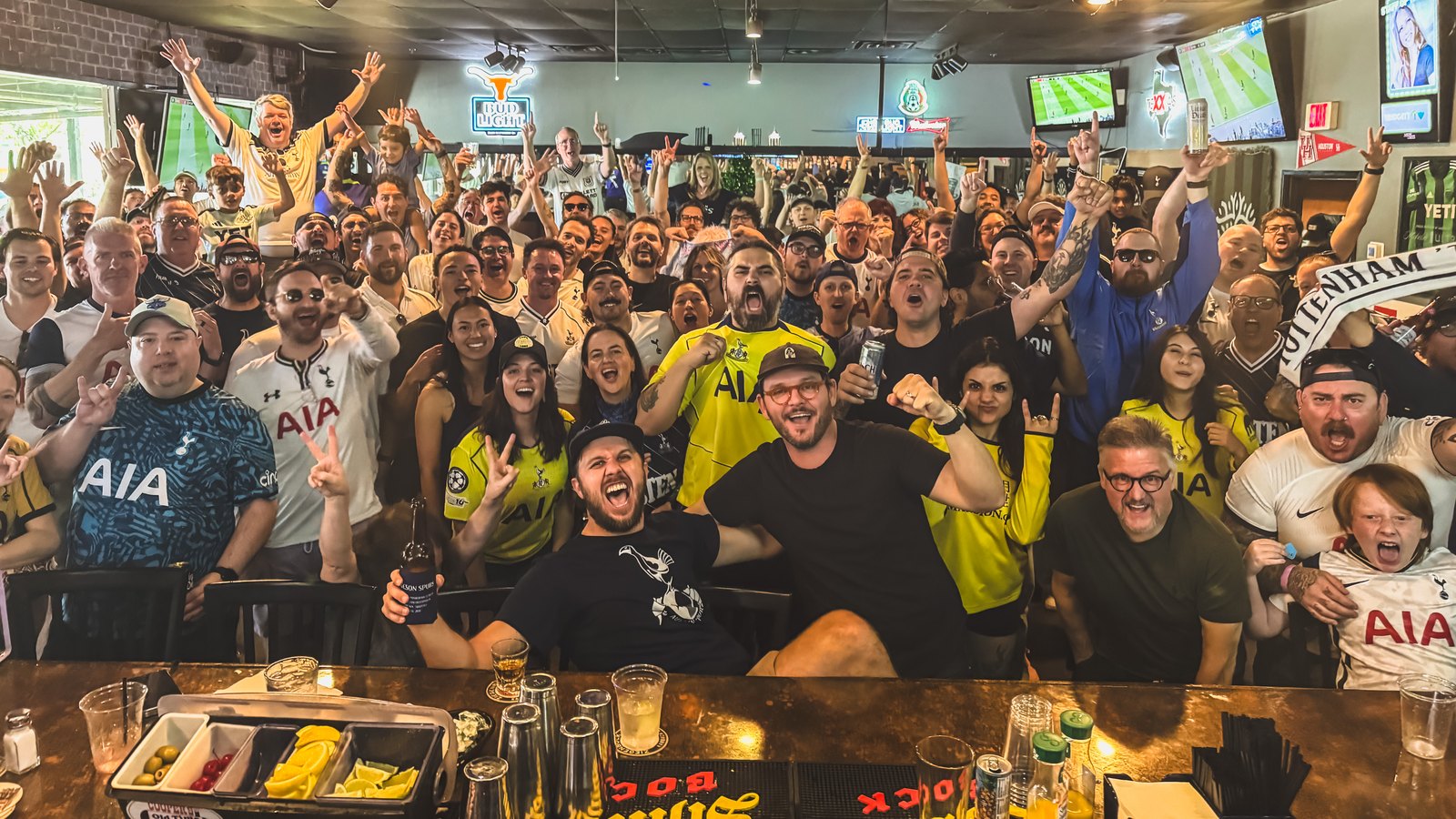 A packed room at Mister Tramps on a matchday: dozens of Austin Spurs supporters in Tottenham shirts and scarves crowded behind the bar, arms raised, with the match on screens overhead.
